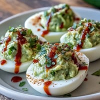Creamy avocado deviled eggs topped with spicy Sriracha drizzle, garnished with fresh cilantro and paprika.